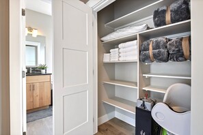 A walk-in closet with shelves for folded clothes, a storage unit with a vacuum, and a door leading to a bathroom.