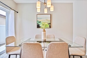 A modern dining area with a glass table, four upholstered chairs, a framed landscape painting, and a pendant light fixture.