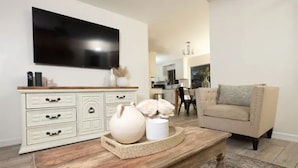 Ein Wohnzimmer mit an der Wand montiertem Fernseher, einem weißen Schrank mit Schubladen, einem beigen Sessel und einem Holztisch mit Dekoration.