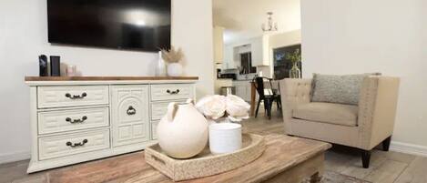 Una sala de estar con un televisor montado en la pared, un mueble blanco con cajones, un sillón color beige y una mesa de centro de madera con objetos decorativos.