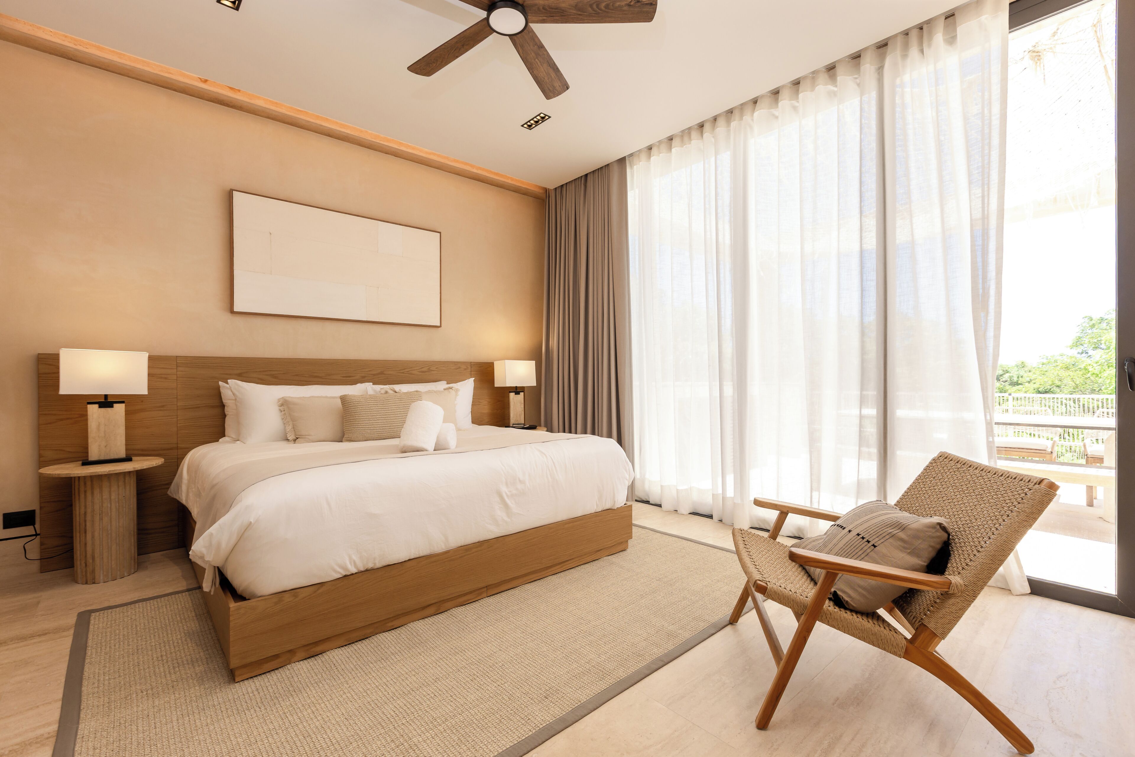 A modern hotel room with a large bed, two bedside tables with lamps, a ceiling fan, and a view of the outdoors through large windows.