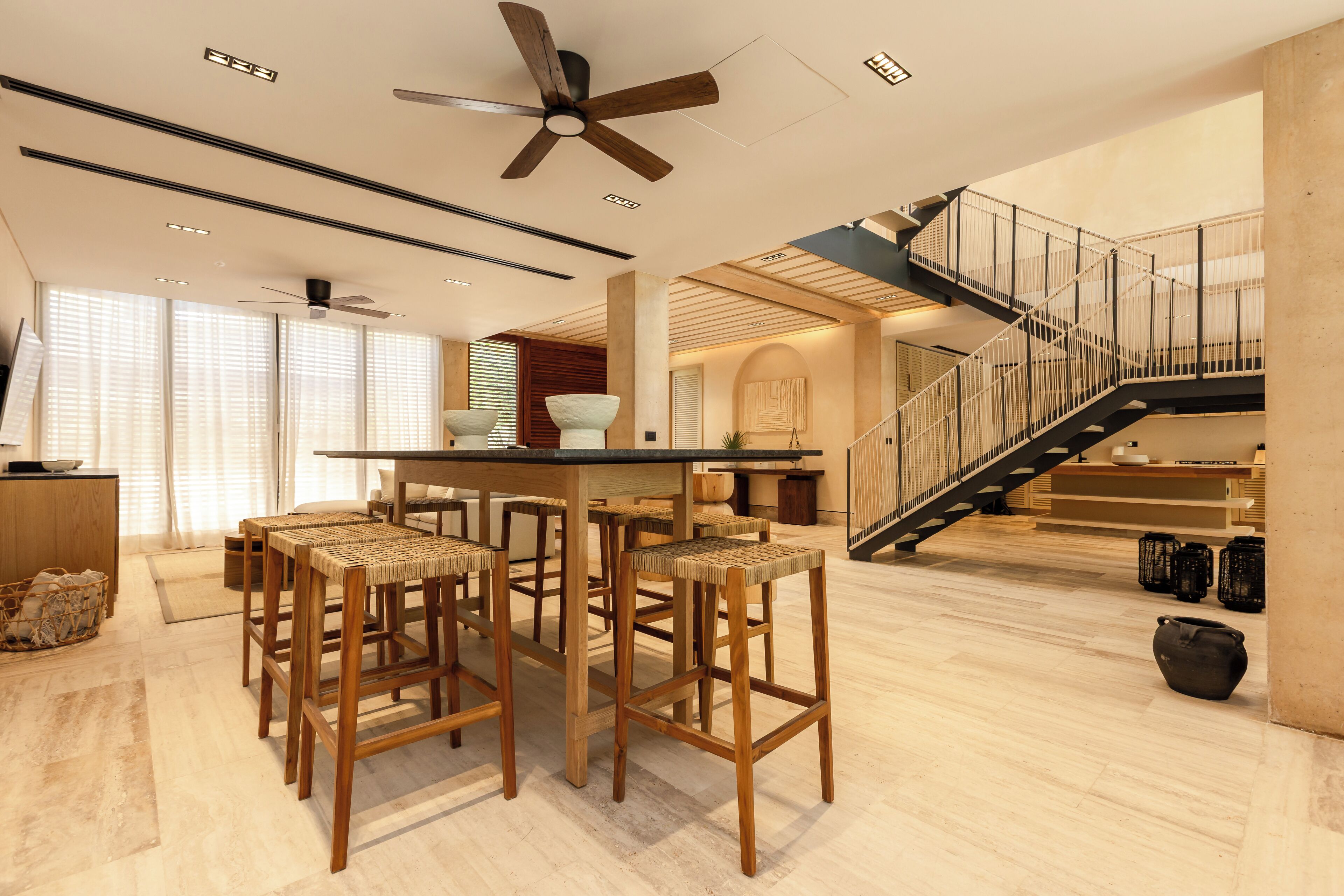Amplia sala de estar con techo alto, una mesa de comedor central con cuatro taburetes, una escalera con barandilla y un gran ventanal con persianas.