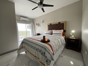 A bedroom with a bed, a ceiling fan, a nightstand, a lamp, and a window with a view of greenery.