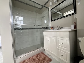 A bathroom with a glass-enclosed shower, white tiled walls, a white vanity with a mirror, and a brown rug.