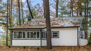 A small, single-story cabin with a metal roof, surrounded by trees and fallen leaves.