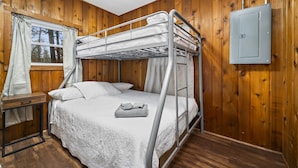A bunk bed room with wooden walls, a window with curtains, a desk, and a power outlet.