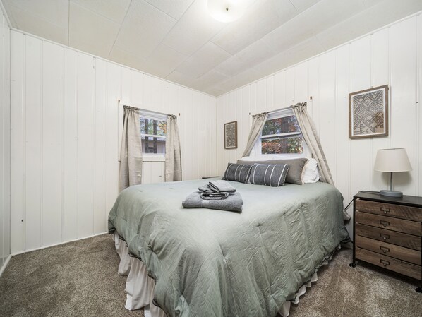 Un dormitorio con cama, cómoda, mesita de noche y dos ventanas con cortinas.