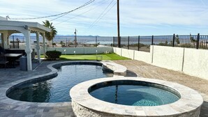 A swimming pool with a curved design, surrounded by stone tiles and a patio area with outdoor furniture. - Peaceful Desert Villa - Lake View, Pool & Spa (Lake Havasu City)