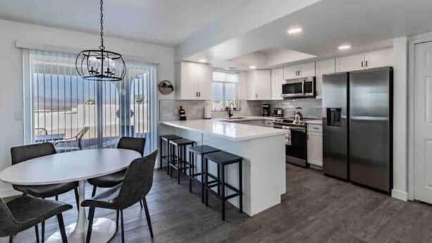 A modern kitchen with a central island, white cabinetry, stainless steel appliances, and a dining area with a round table and chairs. - Peaceful Desert Villa - Lake View, Pool & Spa (Lake Havasu City)
