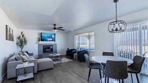 A modern living room with a fireplace, a dining area with chairs, and a large window with blinds. - Peaceful Desert Villa - Lake View, Pool & Spa (Lake Havasu City)