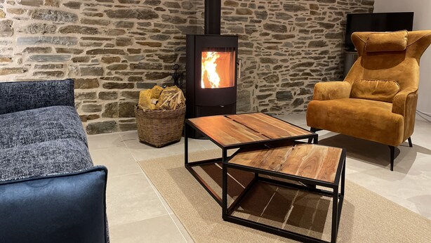 A cozy room with a stone wall, a wood-burning stove, a blue sofa, a mustard-colored armchair, and a woven basket.