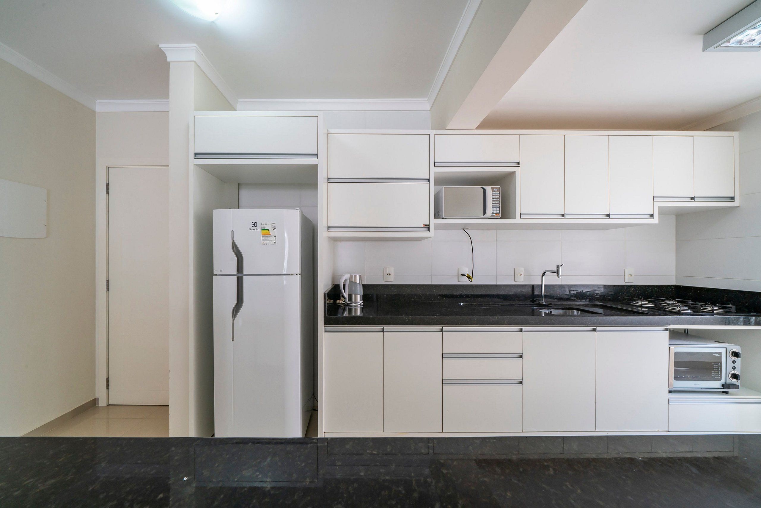 Una cocina moderna con armarios blancos, encimera negra, microondas integrado, refrigerador y tostadora.