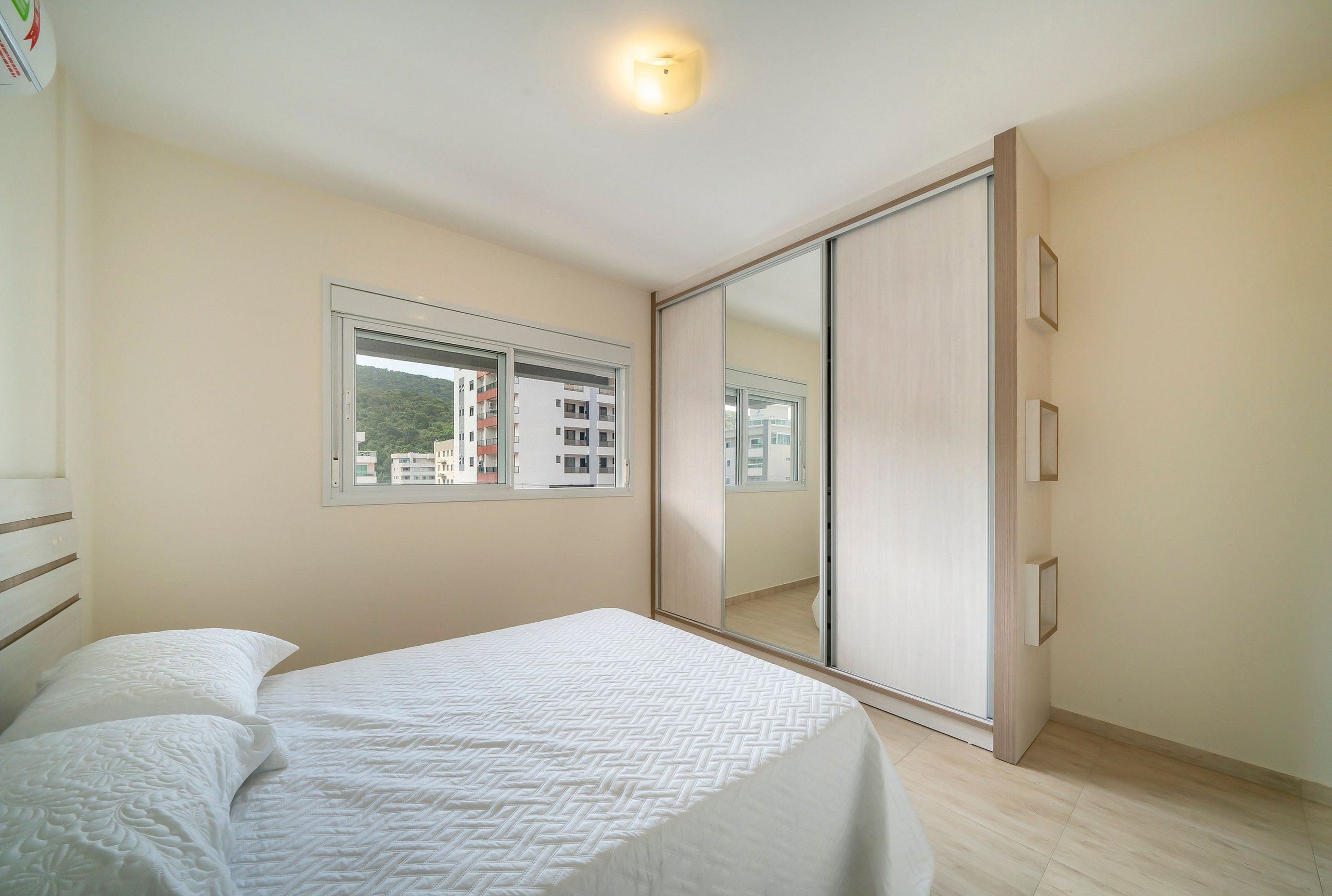 Quarto com cama, janela, porta de correr e vista para prédios.