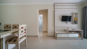 Un salon moderne comprenant un coin repas, un téléviseur fixé au mur et une petite étagère agrémentée d’objets décoratifs.