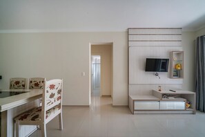 A modern living room with a dining area, a TV mounted on the wall, and a small shelf with decorative items.