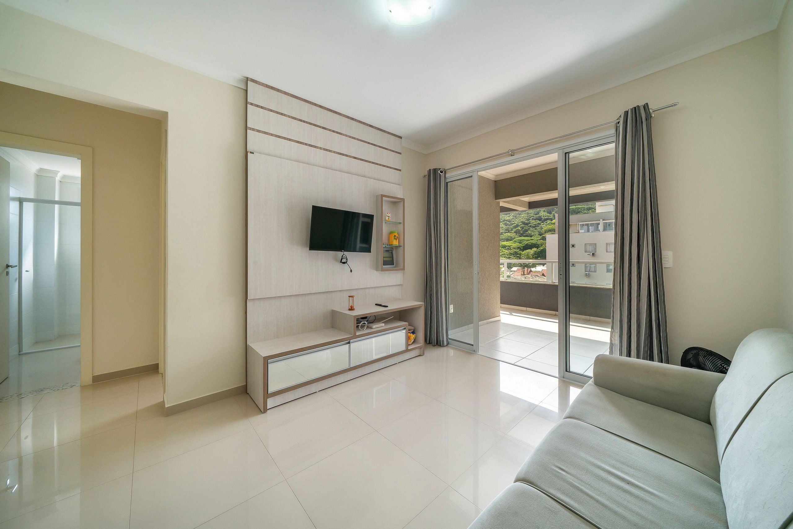 A modern living room with a flat-screen TV mounted on a wall, a sofa, and a sliding glass door leading to a balcony.