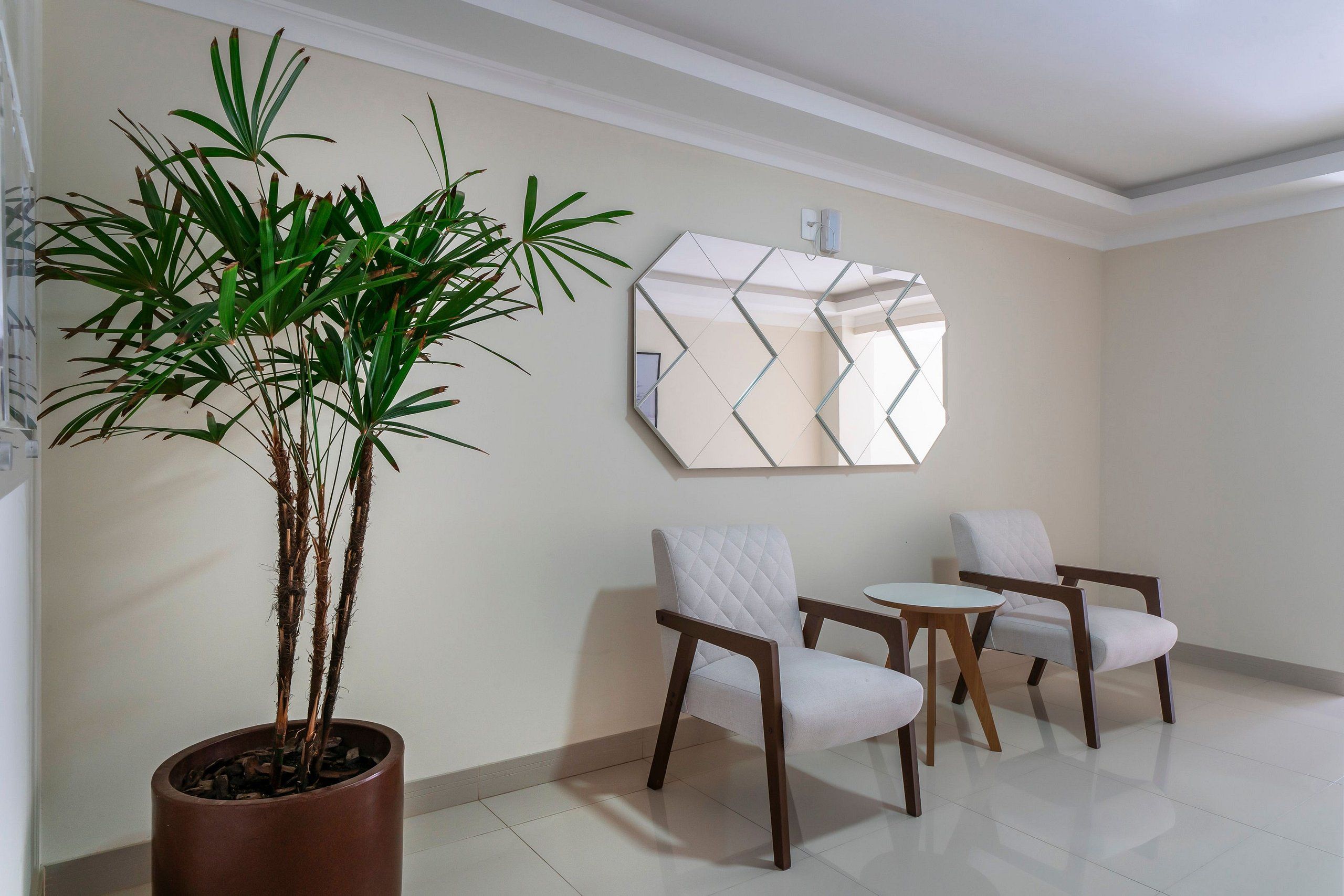 Quarto moderno com planta em vaso, duas poltronas brancas, uma mesinha redonda e um espelho de parede geométrico.