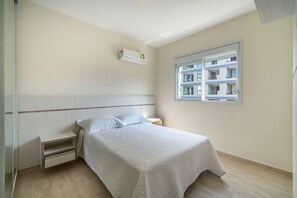 Quarto com cama, mesa de cabeceira, gaveta, janela com vista para edifícios e ar condicionado.