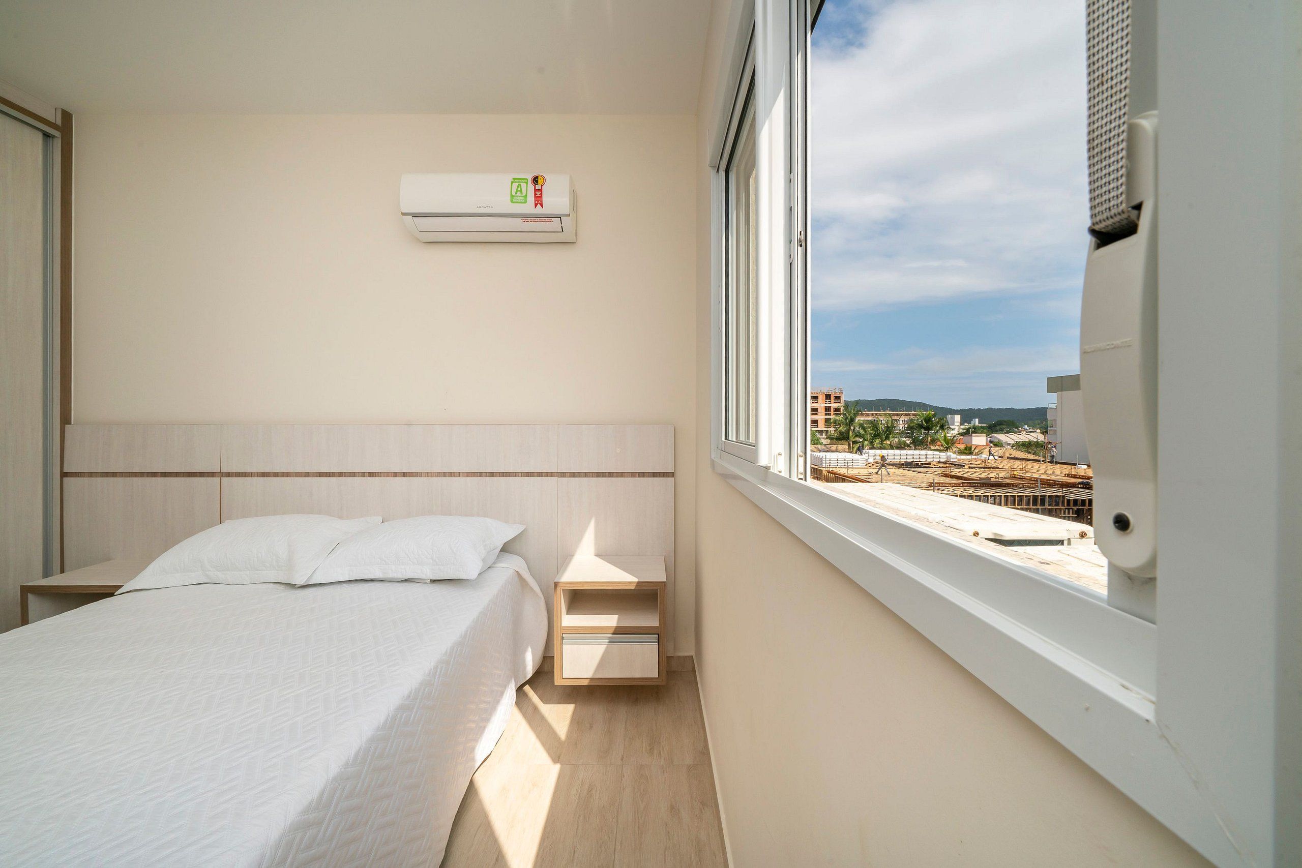 Quarto de hotel com cama, mesa de cabeceira, prateleira de madeira e janela com vista para edifícios e uma obra.