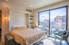Quarto moderno com cama grande, uma cadeira e uma varanda com vista para prédios.