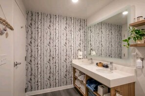 Casa de banho com papel de parede de árvore de bétula, lavatório branco, espelho e prateleira com toalhas e produtos de higiene pessoal.