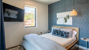A modern bedroom with a bed, a window, a wall-mounted TV, a small table, and a decorative wall piece. - Ski-in/Ski-out Canyon Haus Modern 1 bedroom- Pool, Hot Tub, Gym (Park City)