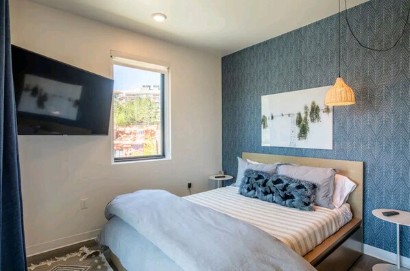 Een moderne slaapkamer met een bed, een raam, een wandtelevisie, een klein tafeltje en een decoratief object aan de muur.