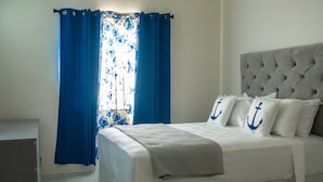 Una cama bien hecha con cabecera gris, ropa de cama blanca y almohadas con anclas azules.