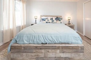 Une chambre à coucher avec une couvre-lit bleu clair, une tête de lit en bois, deux tables de chevet avec des lampes et une fenêtre ornée de rideaux légers.