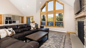 A spacious living room with a large window, a fireplace, and a modern kitchen in the background. - Twin Antler Retreat | Big Sky | Gallatin River (Gallatin Gateway)