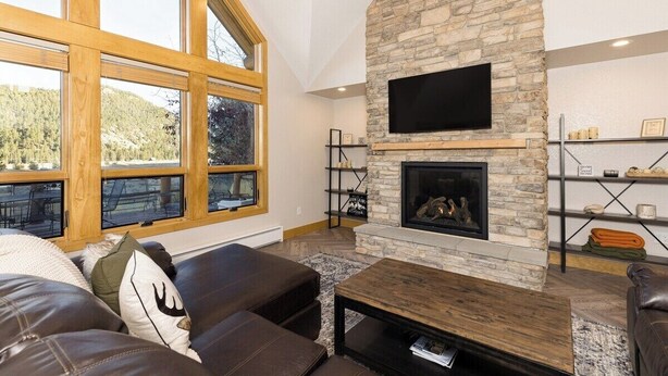 A spacious living room with a stone fireplace, large windows, and a flat-screen TV mounted above it. - Twin Antler Retreat | Big Sky | Gallatin River (Gallatin Gateway)