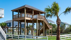 A wooden deck structure with a staircase leading to a balcony, surrounded by palm trees and a white fence.