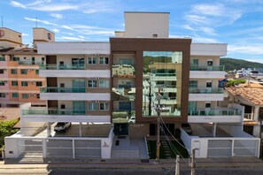Un immeuble moderne à plusieurs étages, doté de balcons, d’une façade centrale en verre et d’un espace de stationnement.