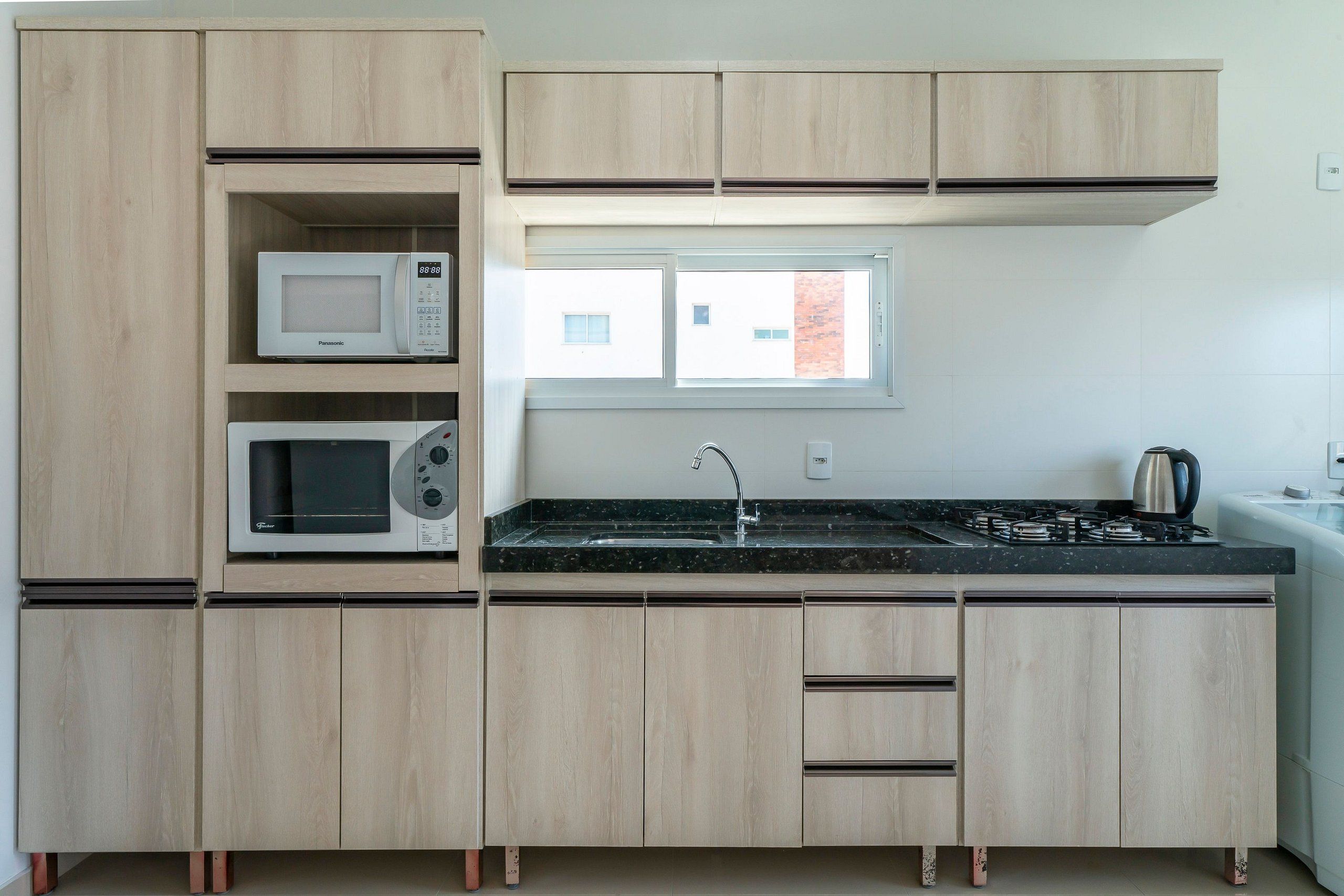 A modern kitchen with wooden cabinets, a black countertop, a stainless steel sink, a gas stove, and two wall-mounted microwaves.