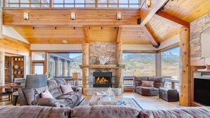 A spacious living room with a stone fireplace, leather sofas, and a large window overlooking a mountainous landscape. - Topaz Estate by Avantstay Luxe Home, Pool + View (Silverthorne)