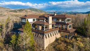 A large, multi-story house with a stone foundation, multiple balconies, and large windows, surrounded by trees and mountains. - Topaz Estate by Avantstay Luxe Home, Pool + View (Silverthorne)