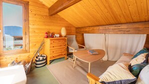 A cozy wooden cabin interior with a wooden ceiling, a window overlooking a mountain, a wooden dresser, a chair, a round table, and a sofa with patterned cushions.
