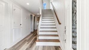 Un escalier aux murs blancs et aux marches en bois, menant à un palier où se trouve un luminaire.