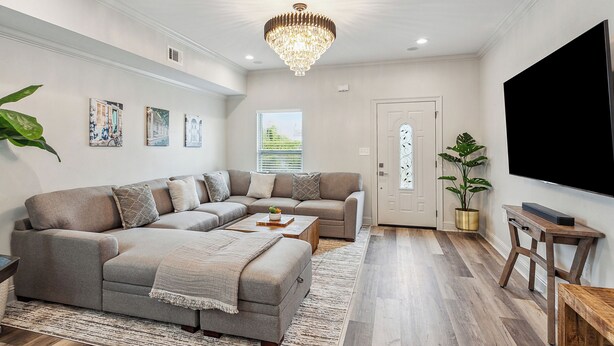Un salon moderne avec un canapé d’angle, une table basse et un lustre. On y trouve une porte avec une fenêtre, une plante et un téléviseur à écran plat fixé au mur.