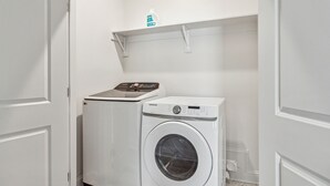 Une petite buanderie équipée d’un lave-linge et d’un sèche-linge à chargement frontal, d’une étagère avec une bouteille et d’une porte dotée d’une poignée.