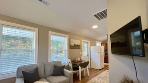 Uma sala de estar com um sofá cinzento, uma mesa de jantar com cadeiras, um frigorífico branco e uma televisão de ecrã plano fixada na parede.