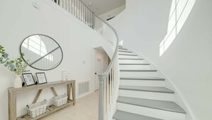 Una escalera con barandillas y escalones blancos, un espejo redondo, una consola con adornos y una planta en maceta.
