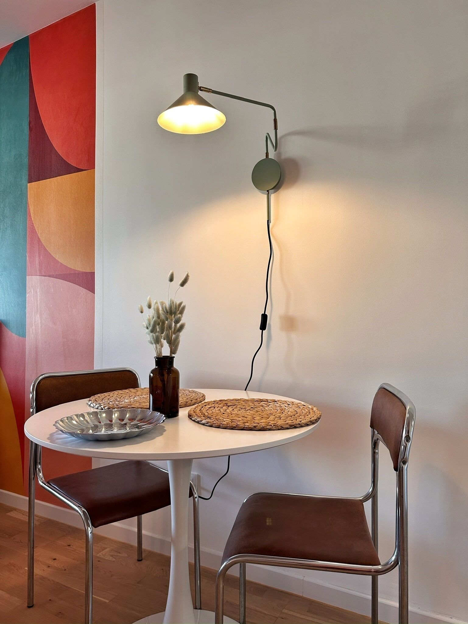 A dining area with a round table, two chairs, a vase with dried flowers, and a modern floor lamp.