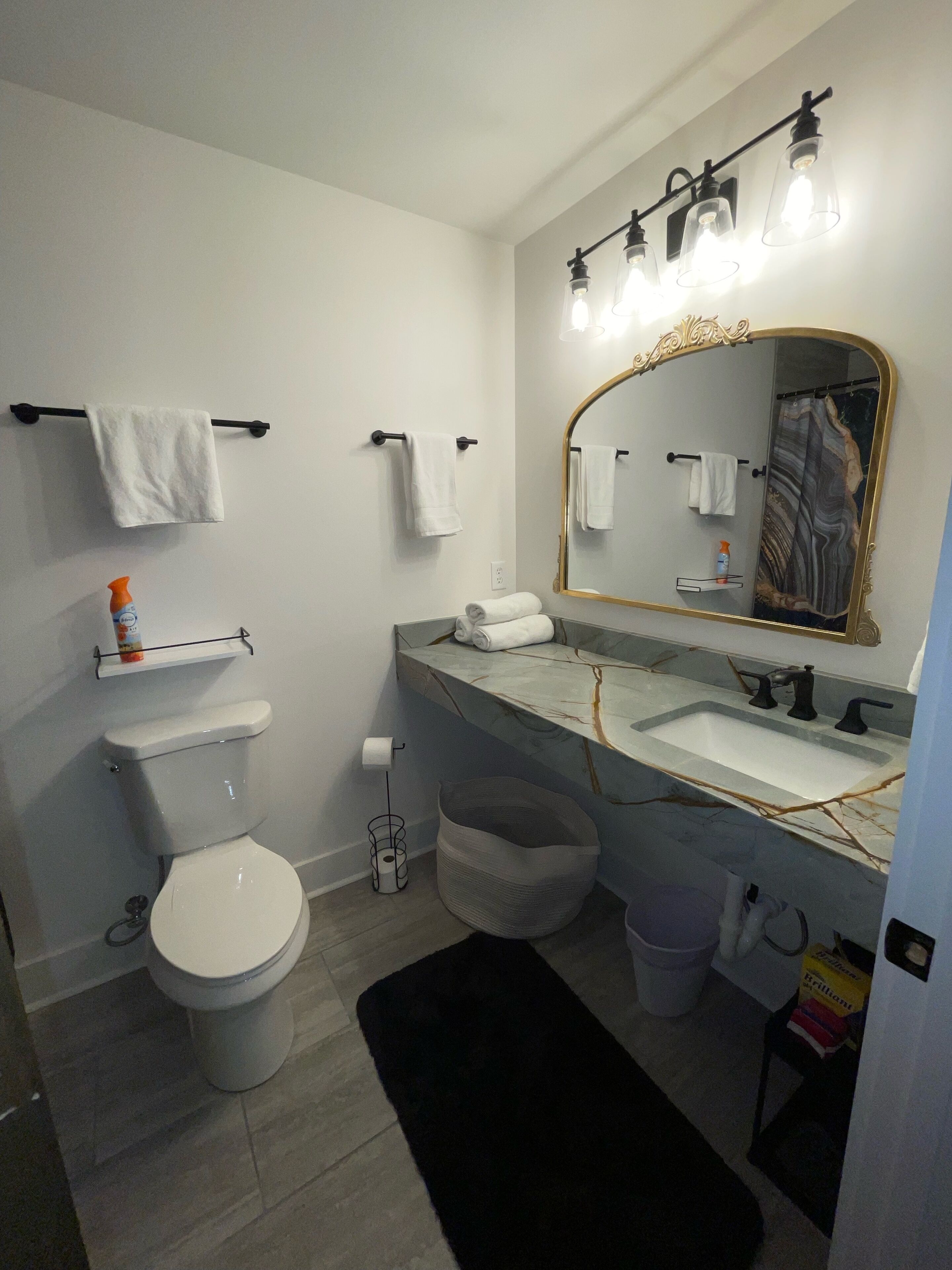 Een moderne badkamer met toilet, wastafel en spiegel. Handdoeken hangen aan een rek, er is een douchegordijn en een klein plankje met toiletartikelen.