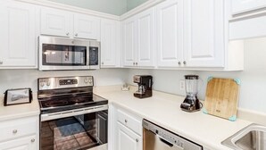 A kitchen with white cabinets, a stainless steel microwave, a stove, a coffee maker, a blender, and a wooden cutting board.