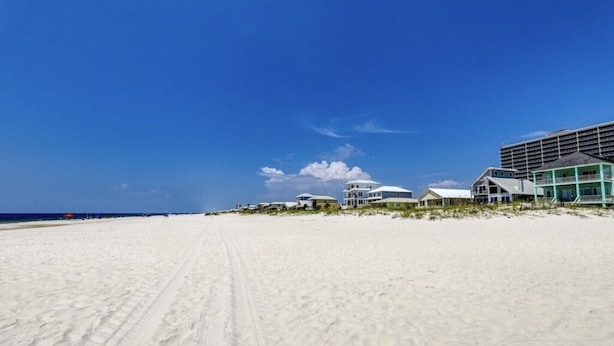 A beach with a clear sky, a sandy path leading to beachfront properties, and a large building with multiple balconies.