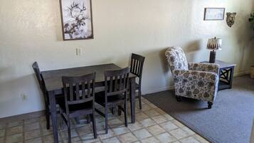 A dining area with a wooden table and chairs, a patterned armchair, a small side table with a lamp, and two framed pictures on the wall.