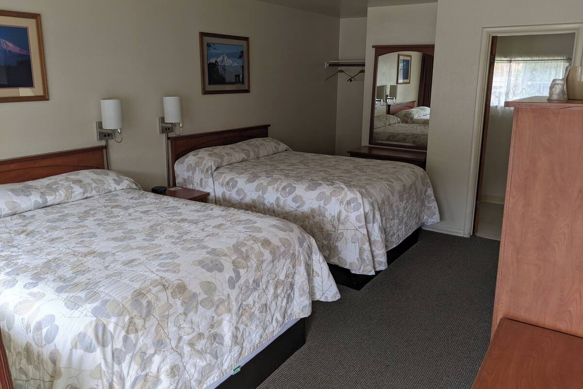 A hotel room with two beds, a wooden headboard, a bedside table with a lamp, a framed picture on the wall, and a mirror reflecting the bed.