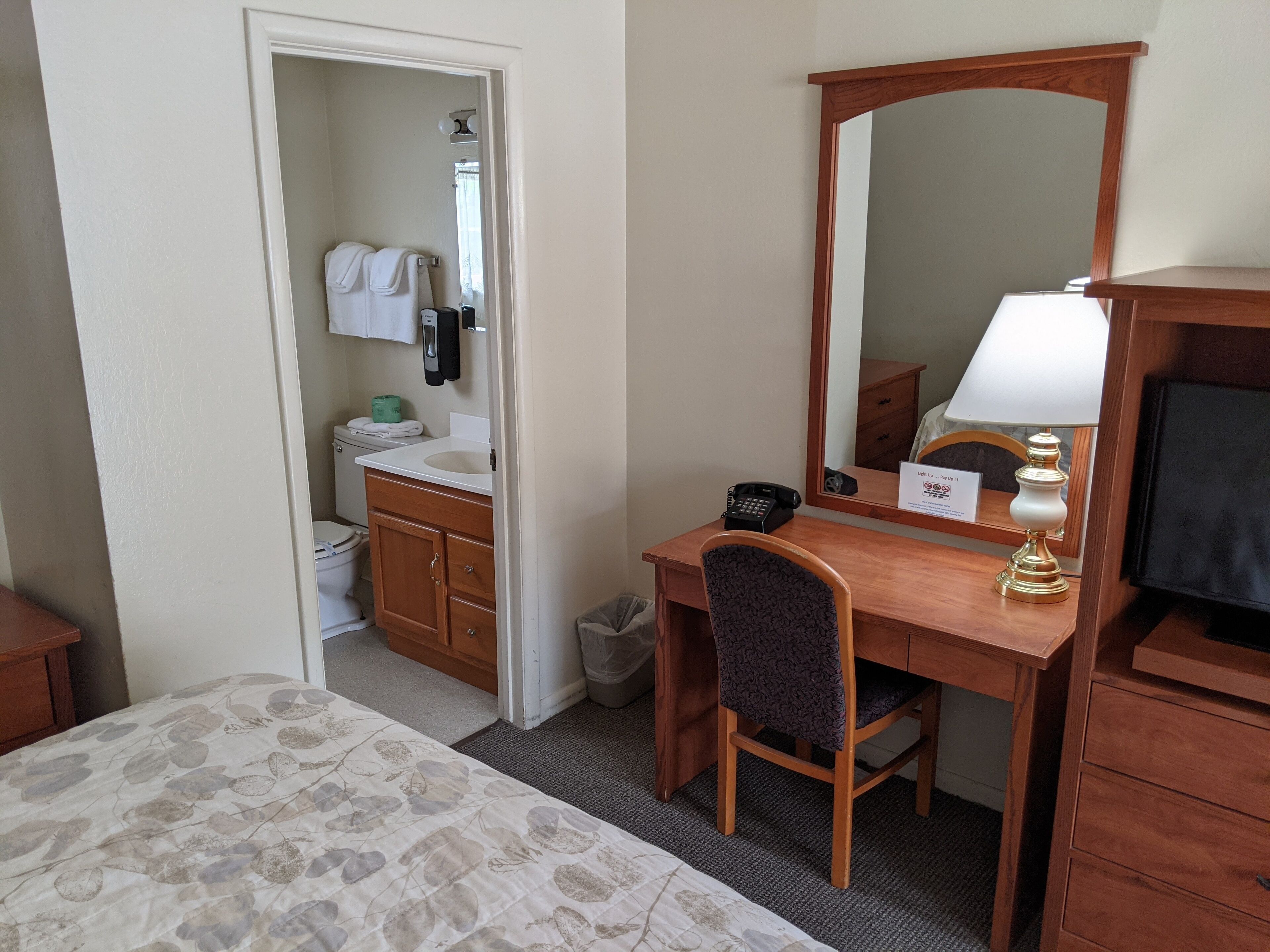 Une chambre d’hôtel comprenant un lit, un bureau avec une chaise, un miroir, une lampe, un téléphone et un petit placard.