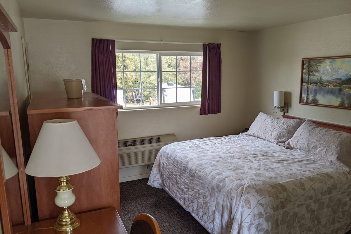 A hotel room with a bed, a desk with a lamp, a window with curtains, a radiator, and a painting on the wall.